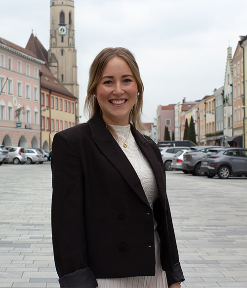 Portrait Foto von Manuela Ganzbeck am Stadtplatz Neuötting mit Kirchturm im Hintergrund.