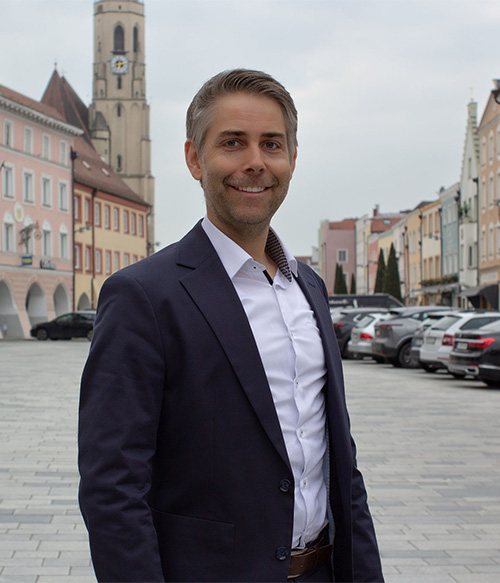 Portrait Foto von Johannes Sigrüner am Stadtplatz Neuötting mit Kirchturm im Hintergrund.